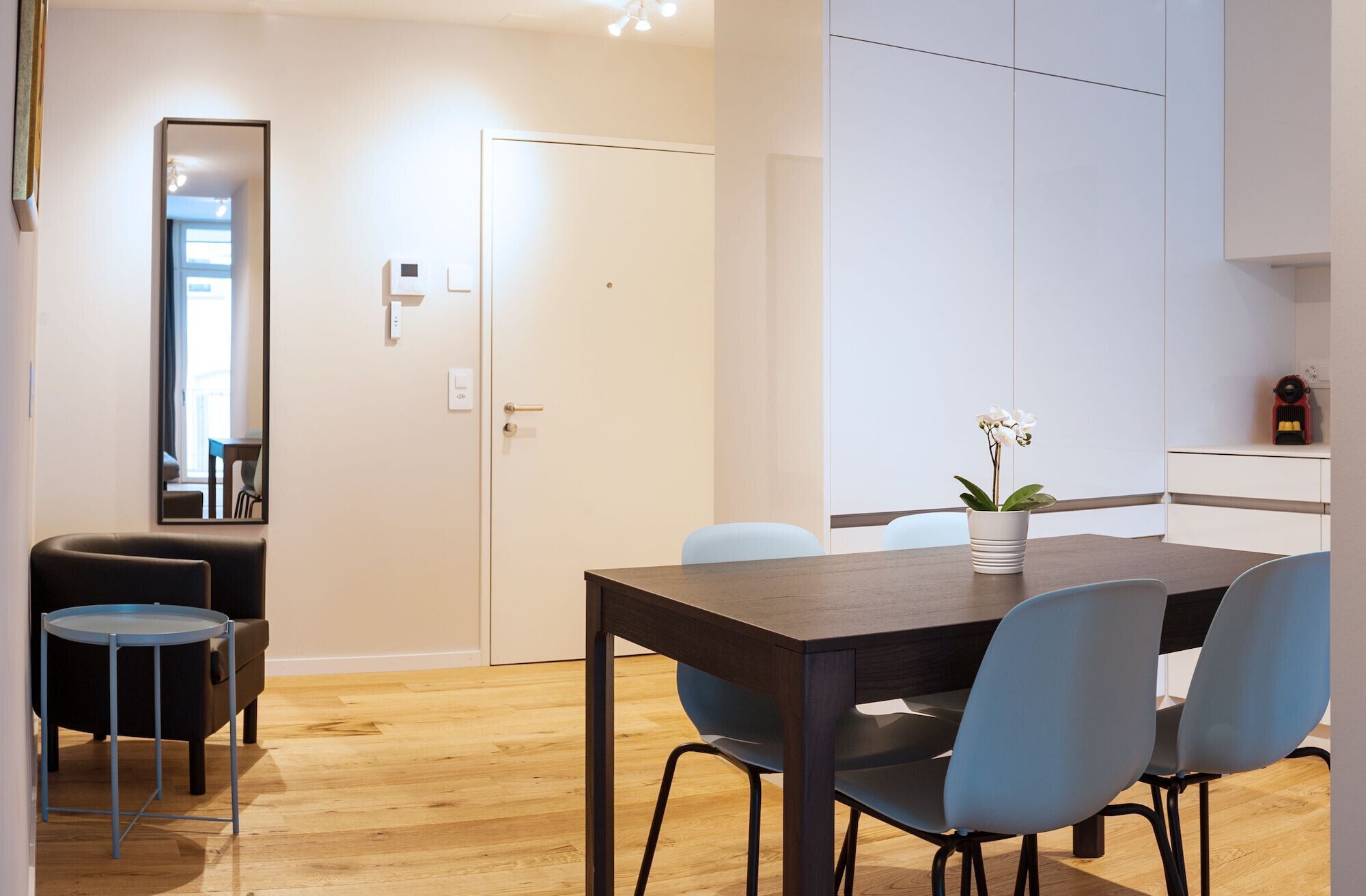 Un comedor moderno con una mesa de madera, cuatro sillas, una orquídea en maceta pequeña y un espejo en la pared.