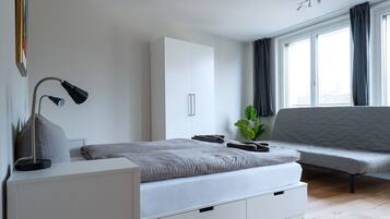 Ein modernes Schlafzimmer mit Bett, Nachttisch, Lampe, Kleiderschrank, Sofa und einem Fenster mit Vorhängen.