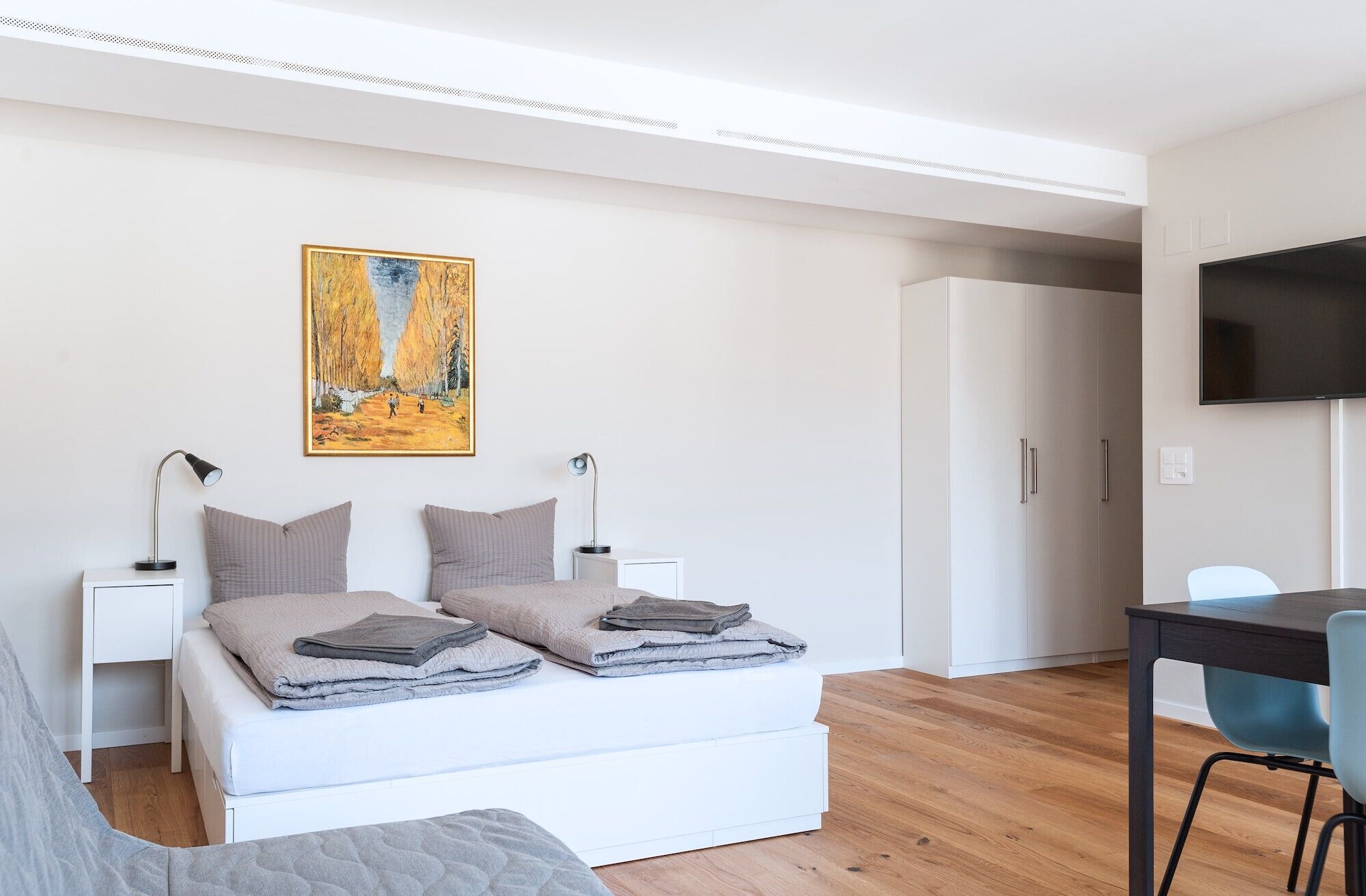 A modern bedroom with a bed, bedside lamps, a painting, a wardrobe, and a TV.