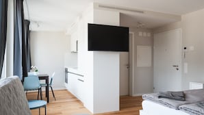 Un espacio habitable moderno y compacto con kitchenette, televisor de pantalla plana sobre la encimera, comedor con mesa y sillas, y cama con manta gris.