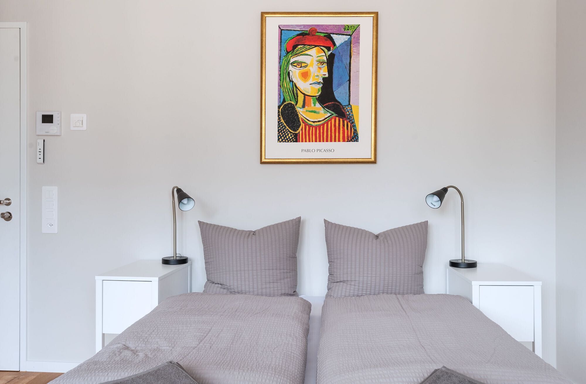 A bedroom with two beds, bedside tables, lamps, and a framed artwork on the wall.
