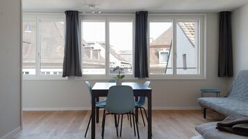 Ein Zimmer mit Esstisch, Stühlen, einer Couch und einem großen Fenster mit Vorhängen.
