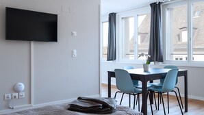 Una habitación moderna con televisión de pantalla plana en la pared, una mesa de comedor con sillas, una cama con una manta doblada y una ventana con cortinas.