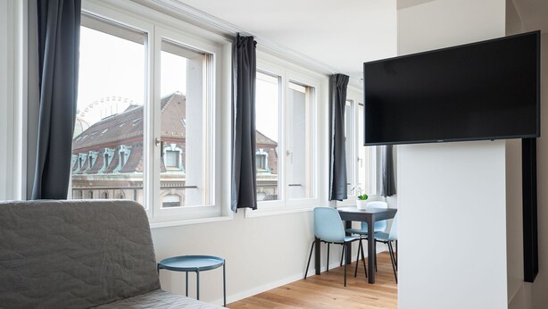 Una sala de estar moderna con un sofá gris, un comedor con una mesa y sillas, ventanas amplias con cortinas y un televisor montado.