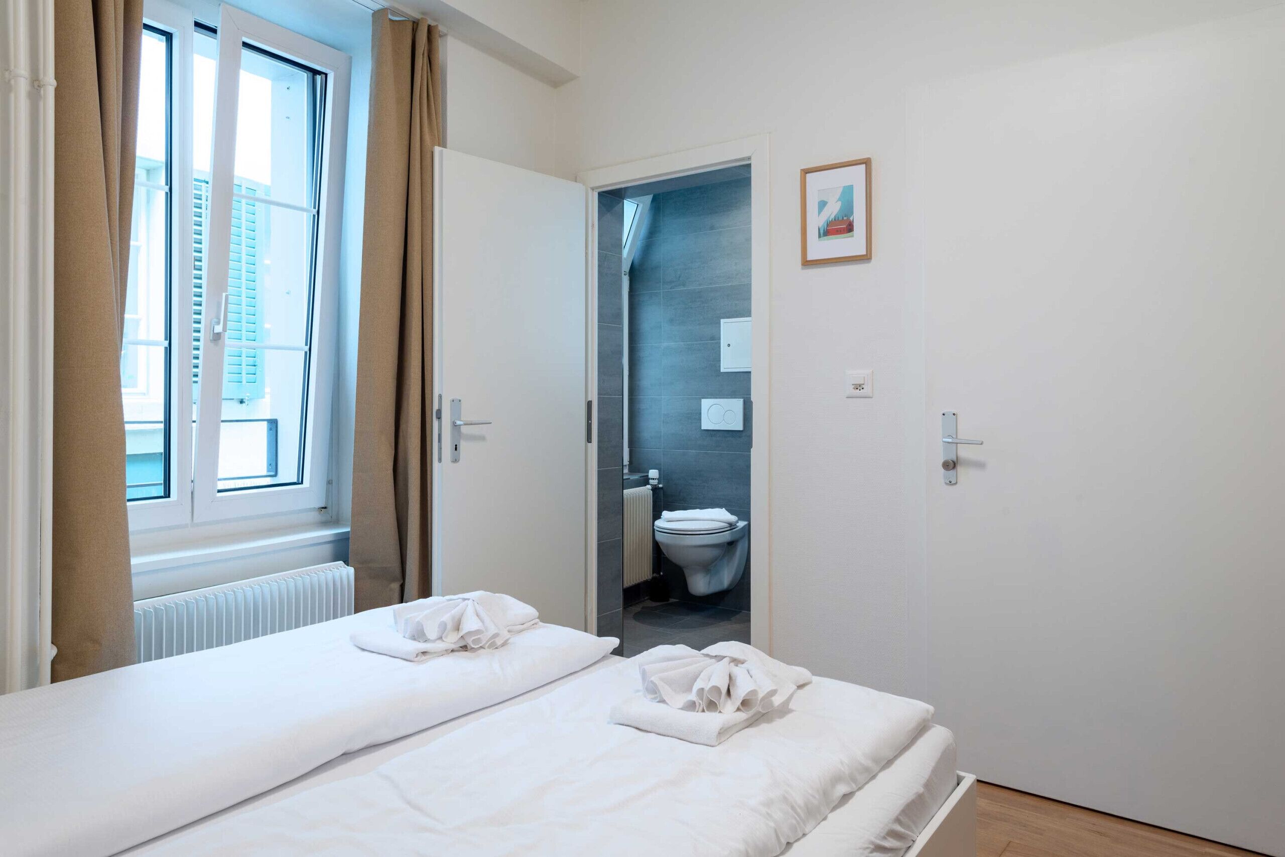 A hotel room with a bed, a window with curtains, a door leading to a bathroom, and a framed picture on the wall.