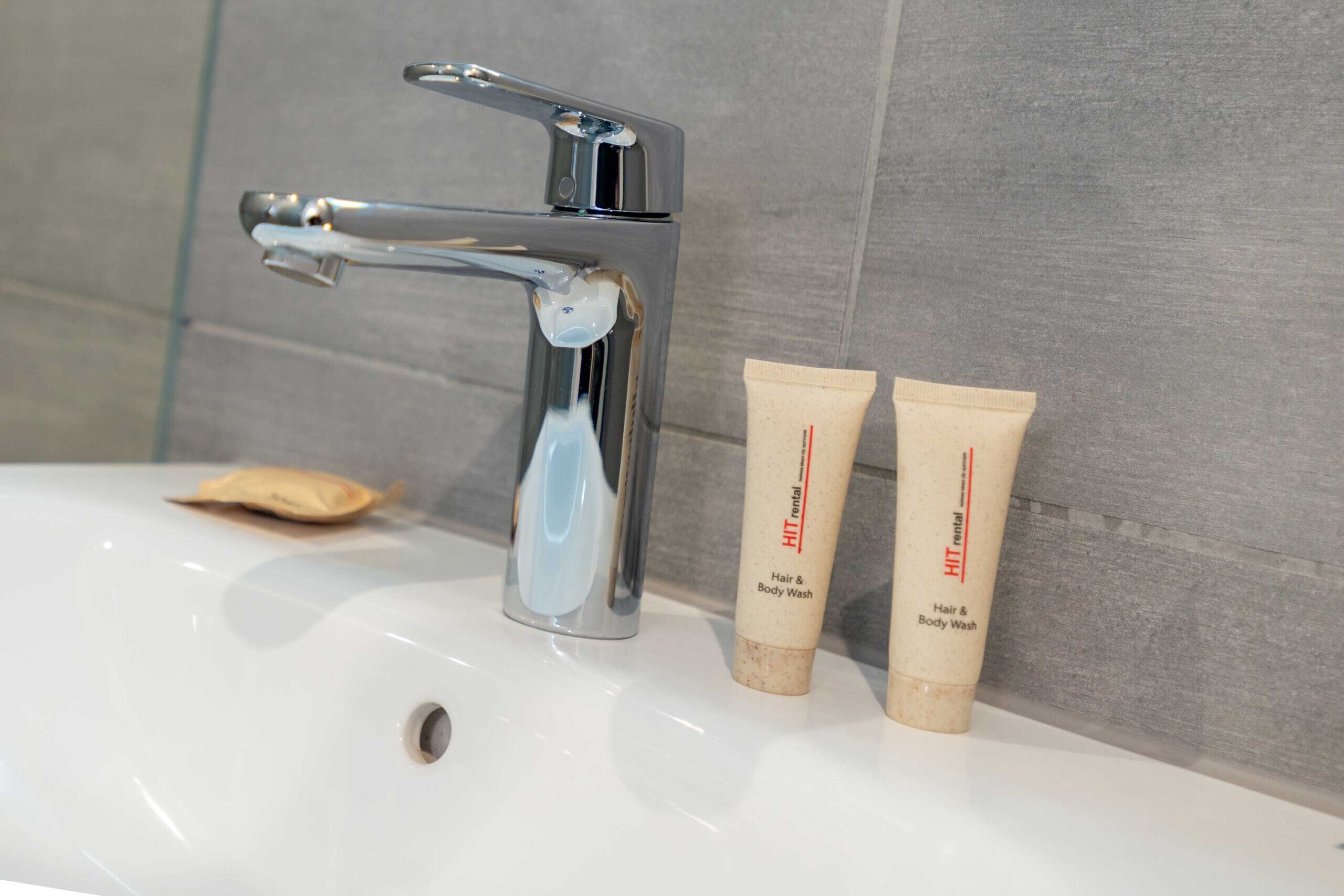 Un lavamanos con grifo de cromo, dos tubos de gel y champú para cabello y cuerpo de HIT alquiler, y una barra de jabón.