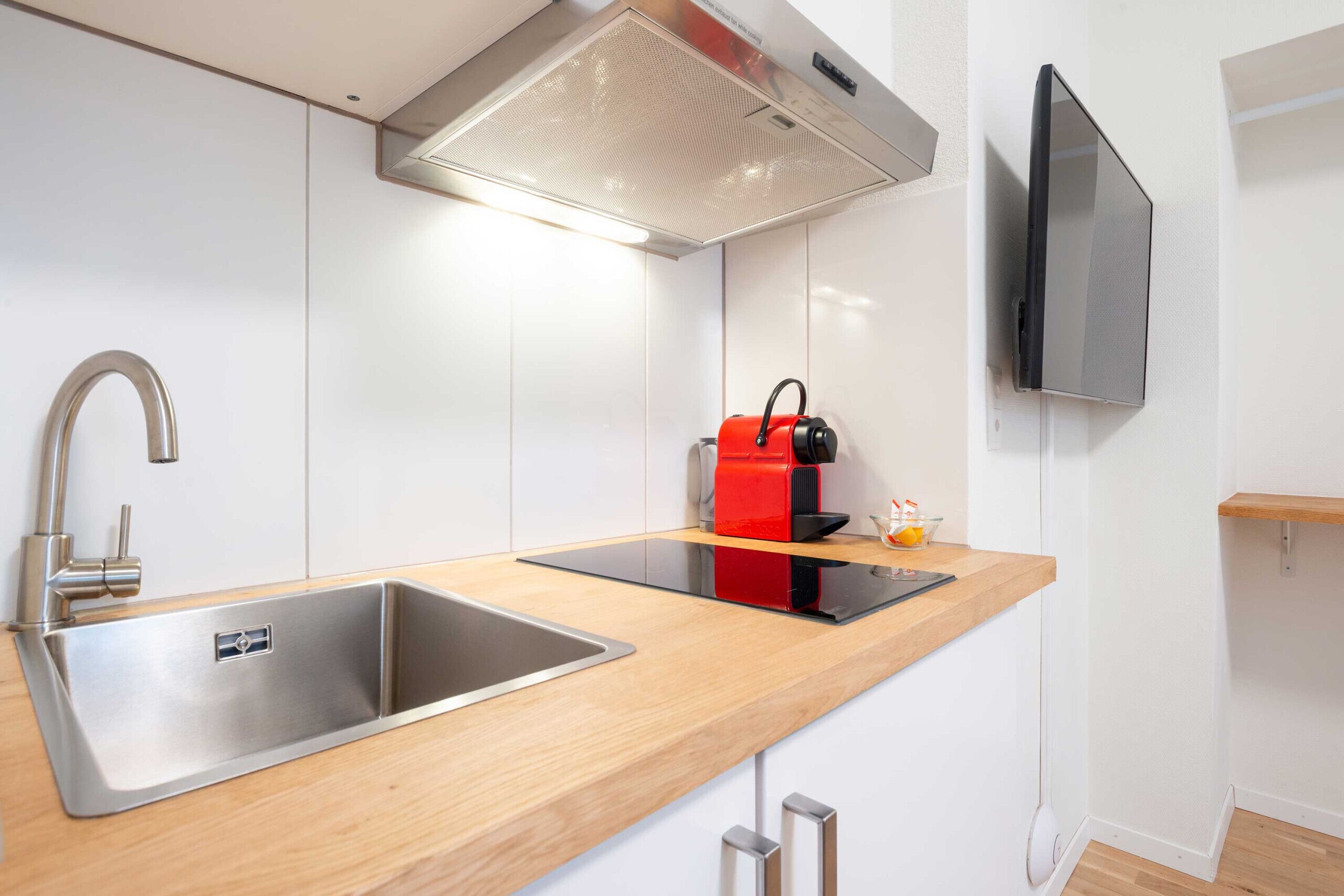 Uma cozinha moderna com pias de aço inoxidável, uma cafeteira vermelha e uma TV de tela plana na parede.