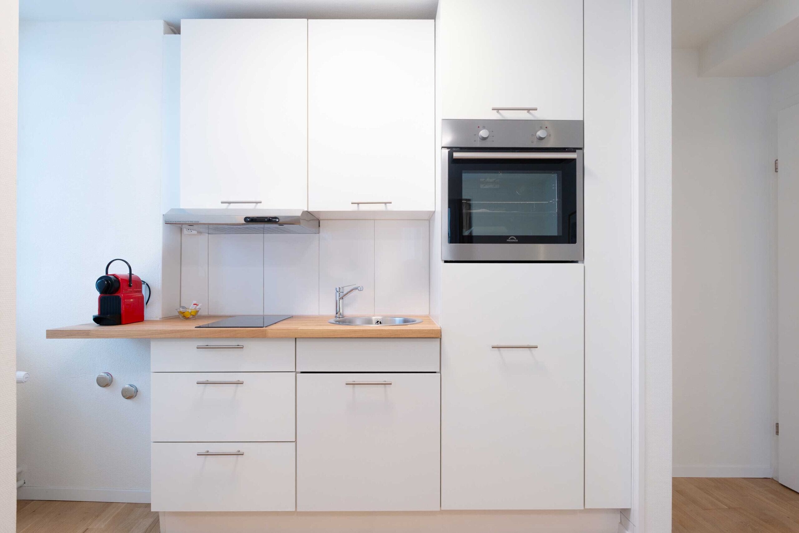 Une cuisine compacte avec des meubles blancs, un four en acier inoxydable, un plan de travail en bois et un évier.