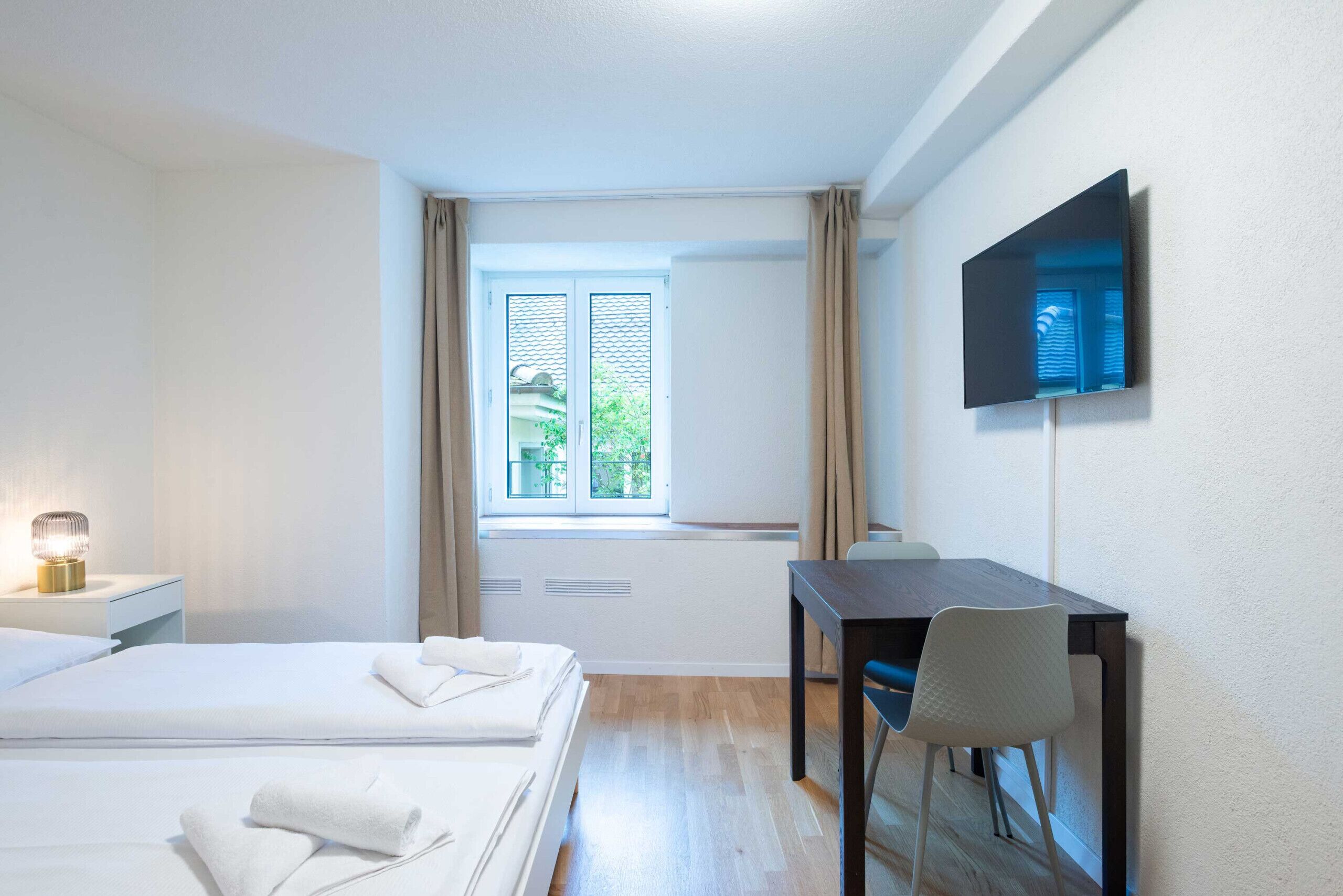 Ein Hotelzimmer mit Bett, Nachttisch mit Lampe, Schreibtisch mit Stuhl, Wand-Fernseher und einem Vorhangfenster.