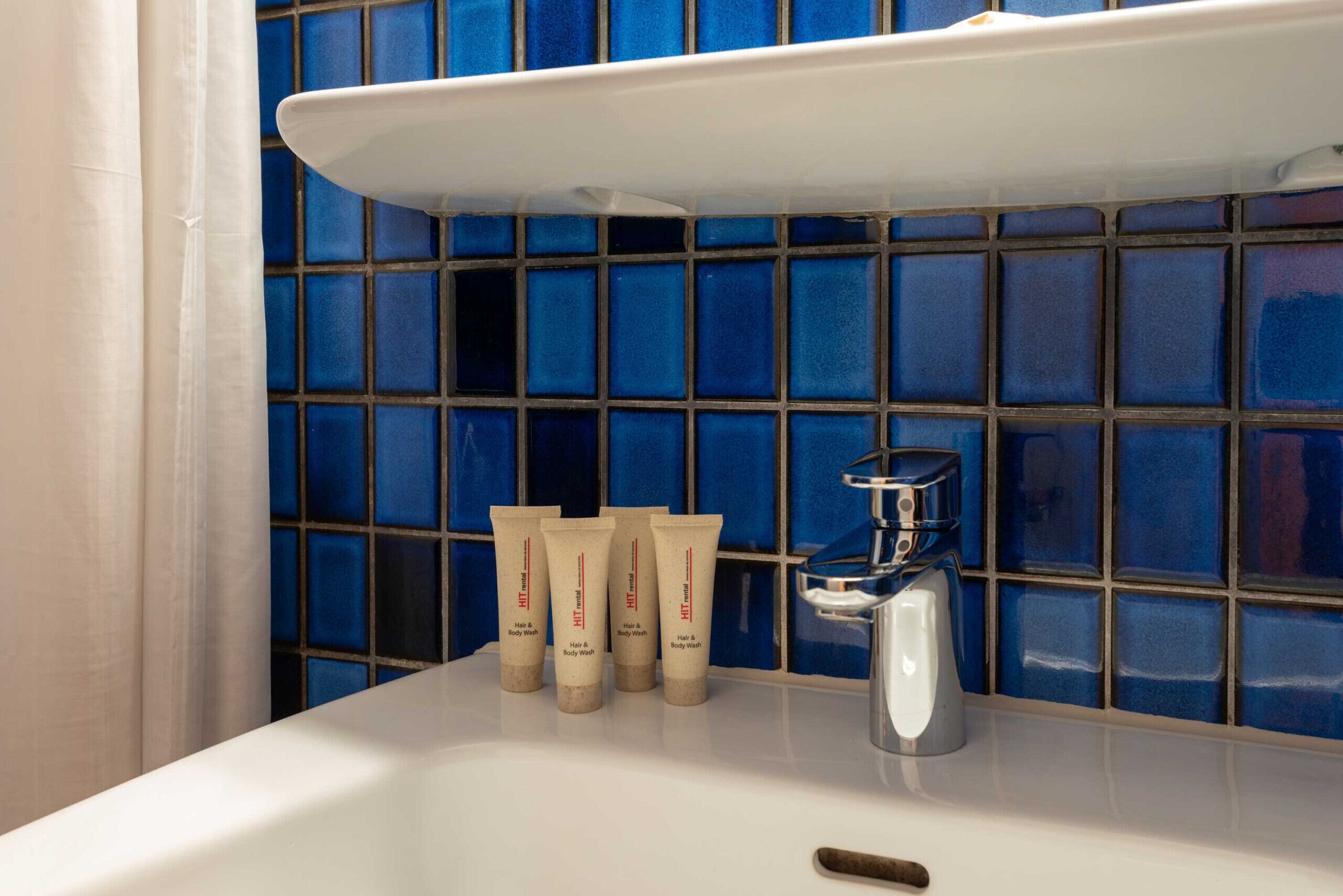 A bathroom with a white sink, blue-tiled wall, and three tubes of hair and body wash.