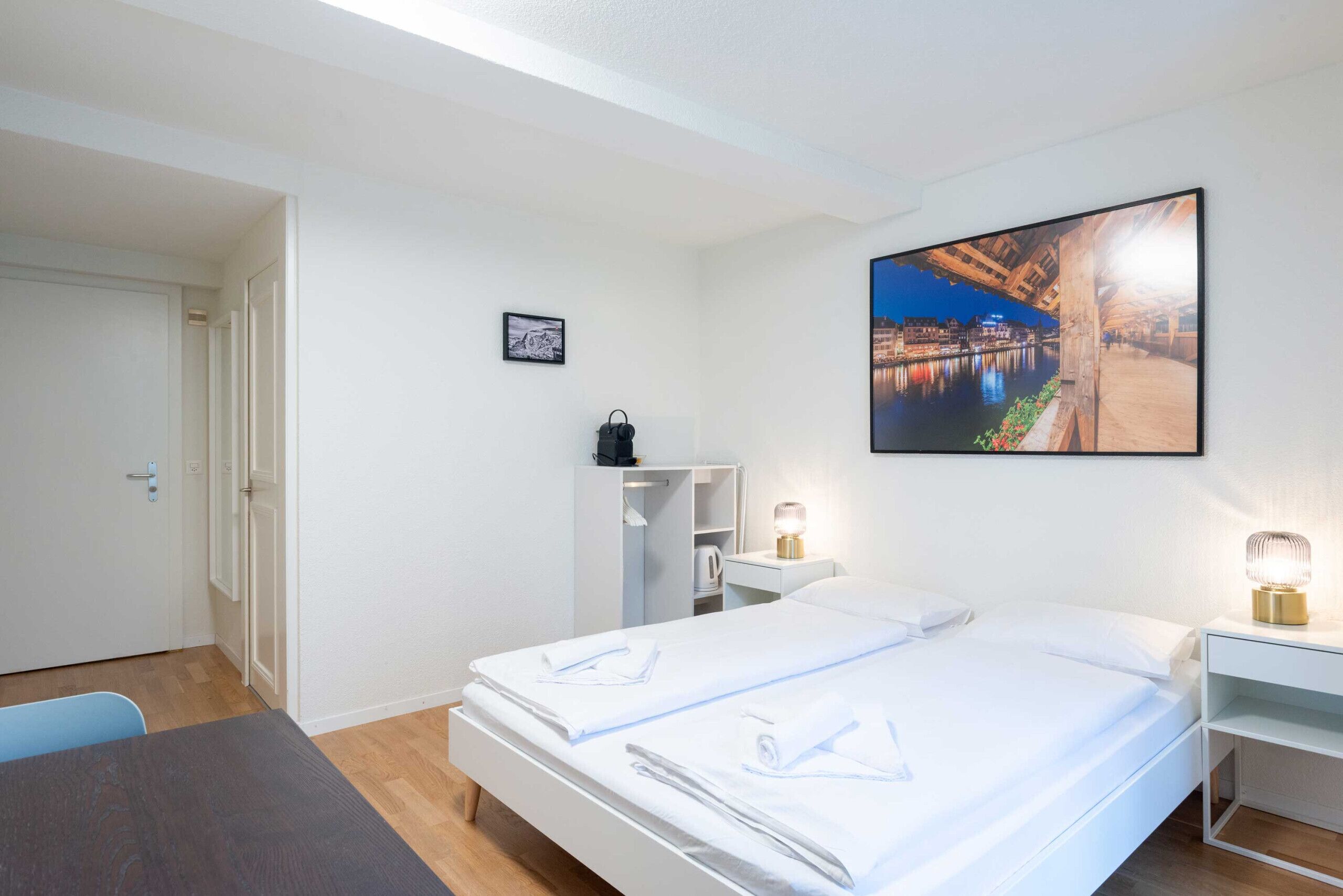 A hotel room with a bed, a desk, a chair, a wardrobe, and a picture of a cityscape.