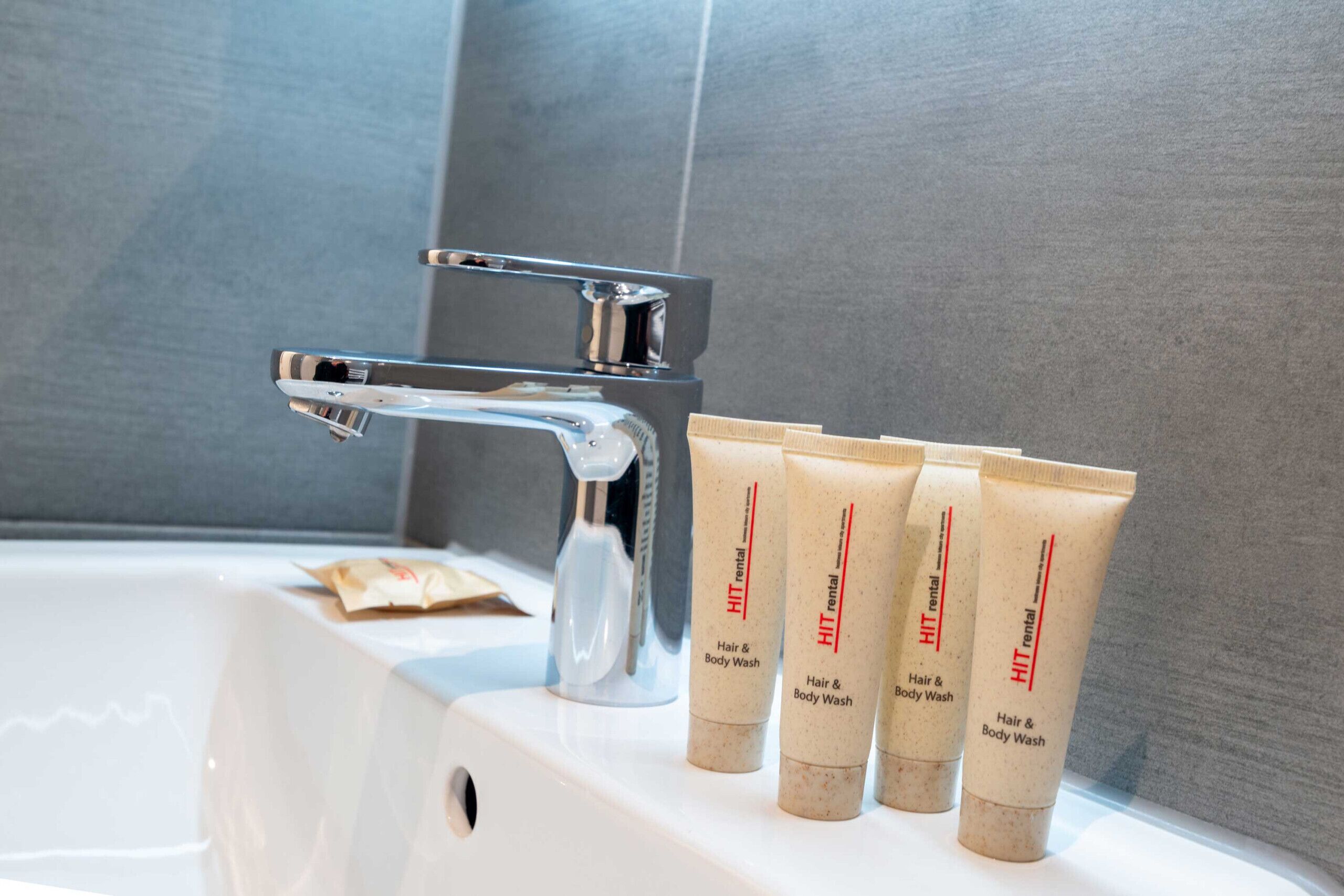 Ein Badezimmer mit einem weißen Waschbecken, einem chromfarbenen Wasserhahn und drei beige gefärbten Flaschen mit dem Aufdruck 'Hair & Body Wash' auf einer weißen Arbeitsplatte.