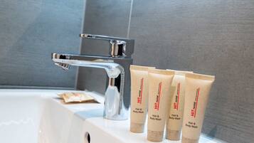Ein Badezimmer mit einem weißen Waschbecken, einem chromfarbenen Wasserhahn und drei beige gefärbten Flaschen mit dem Aufdruck 'Hair & Body Wash' auf einer weißen Arbeitsplatte.