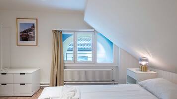 Un dormitorio con cama, mesita de noche con lámpara, ventana con cortinas, radiador y un cuadro en la pared.