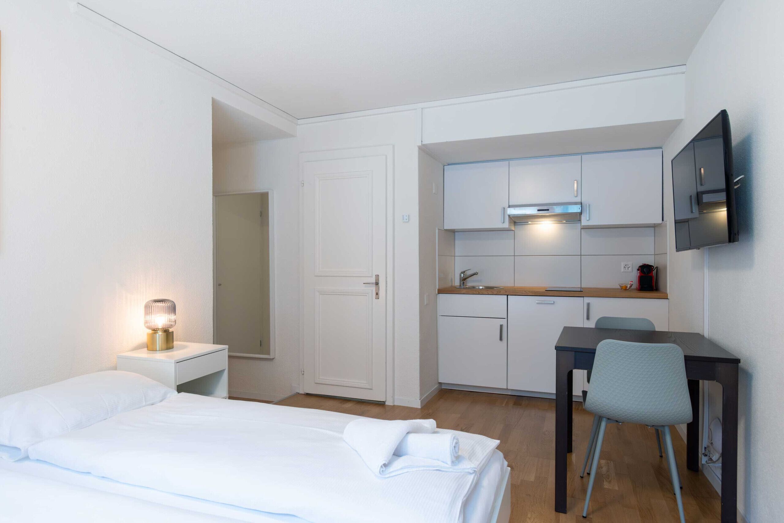 A compact hotel room with a bed, bedside table, lamp, kitchenette, and a TV mounted on the wall.