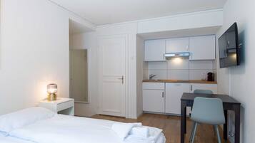 A compact hotel room with a bed, bedside table, lamp, kitchenette, and a TV mounted on the wall.