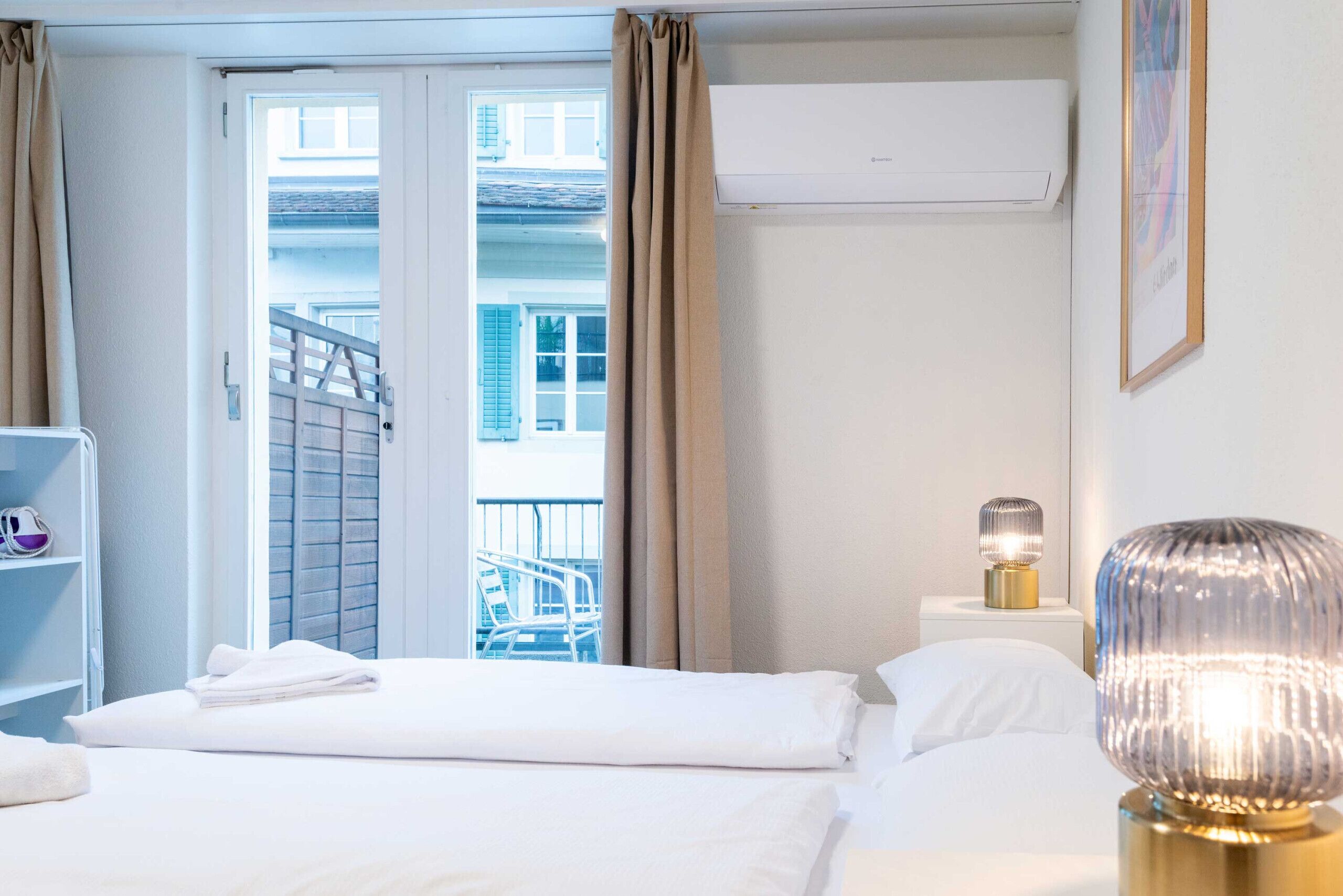 Una camera d'albergo con un letto, un comodino con una lampada, una finestra con tende e un condizionatore d'aria.