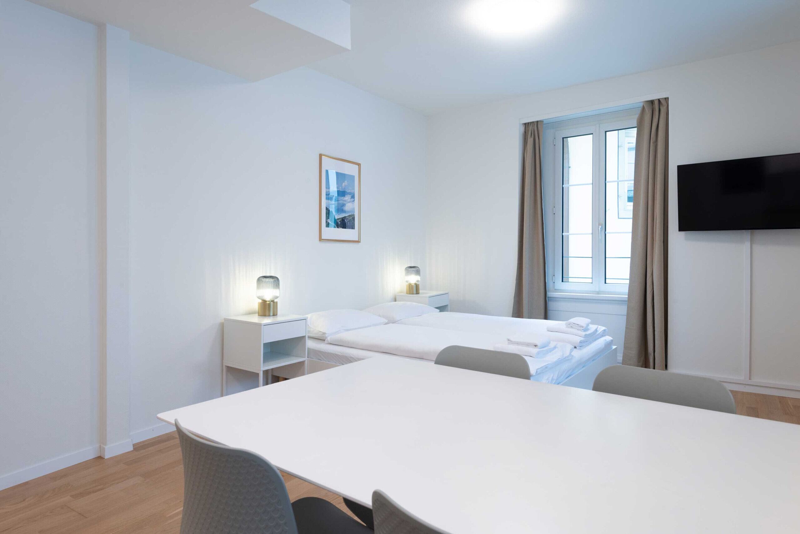 Habitación de hotel con cama, escritorio, silla, televisión, ventana con cortinas y un cuadro en la pared.
