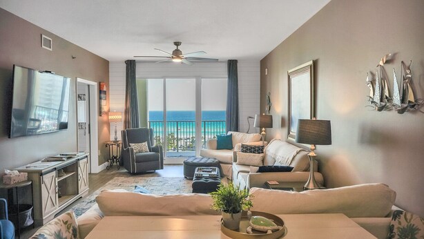 A spacious living room with a large TV, comfortable seating, a balcony with a view of the ocean, and a ceiling fan.