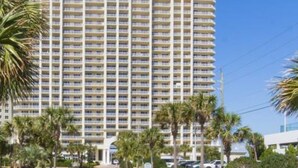 A high-rise hotel with multiple balconies, surrounded by palm trees and a clear blue sky.