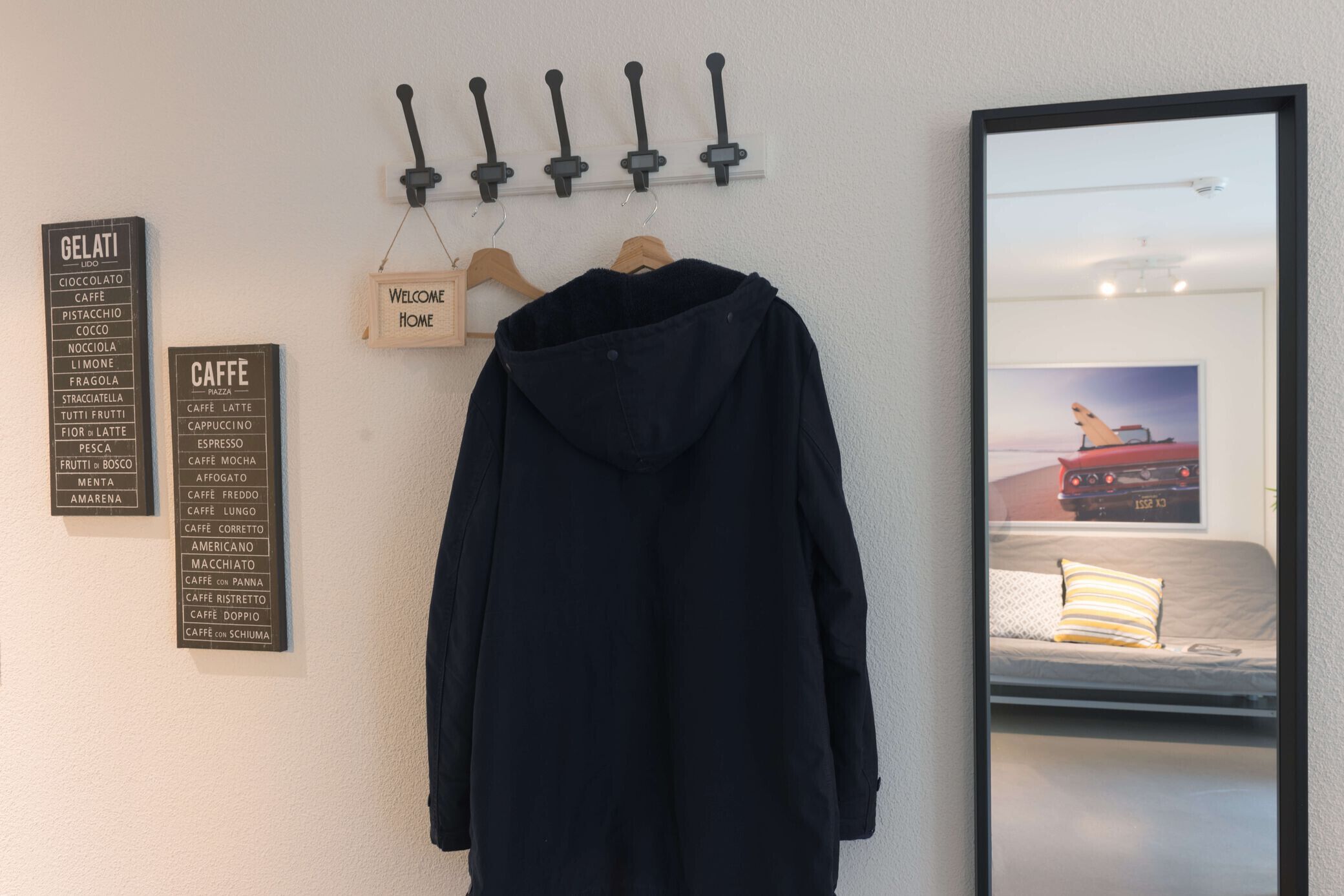 A black jacket hanging on a wooden hanger, a wall-mounted sign listing various coffee types, and a framed picture of a car on a bed.