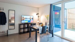 A modern living room with a flat-screen TV, a dining table set for two, and a large sliding glass door leading to an outdoor balcony. - Hitrental Allmend Comfort Apartments (Lucerne)