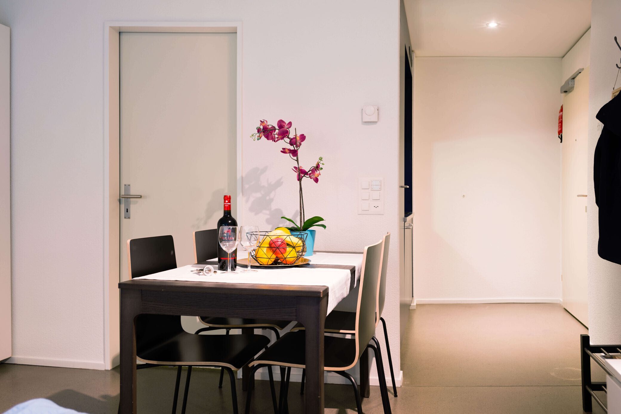 A dining area with a table set for two, a vase with flowers, and a bottle of wine.