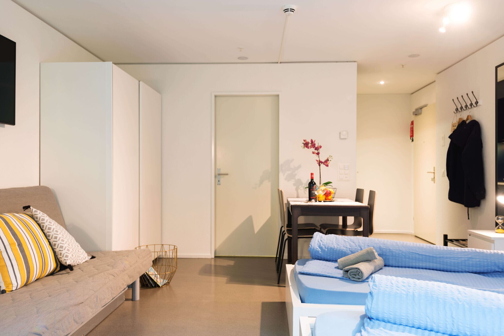 Una habitación moderna y bien iluminada, con sofá, cama, mesa de comedor con sillas y una pequeña cocina.