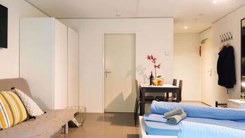 Una habitación moderna y bien iluminada, con sofá, cama, mesa de comedor con sillas y una pequeña cocina.