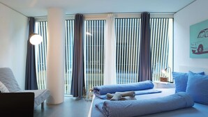 Ein modernes Hotelzimmer mit einem großen Bett, einer Sitzecke mit einem Sessel und einem großen Fenster mit Jalousien.