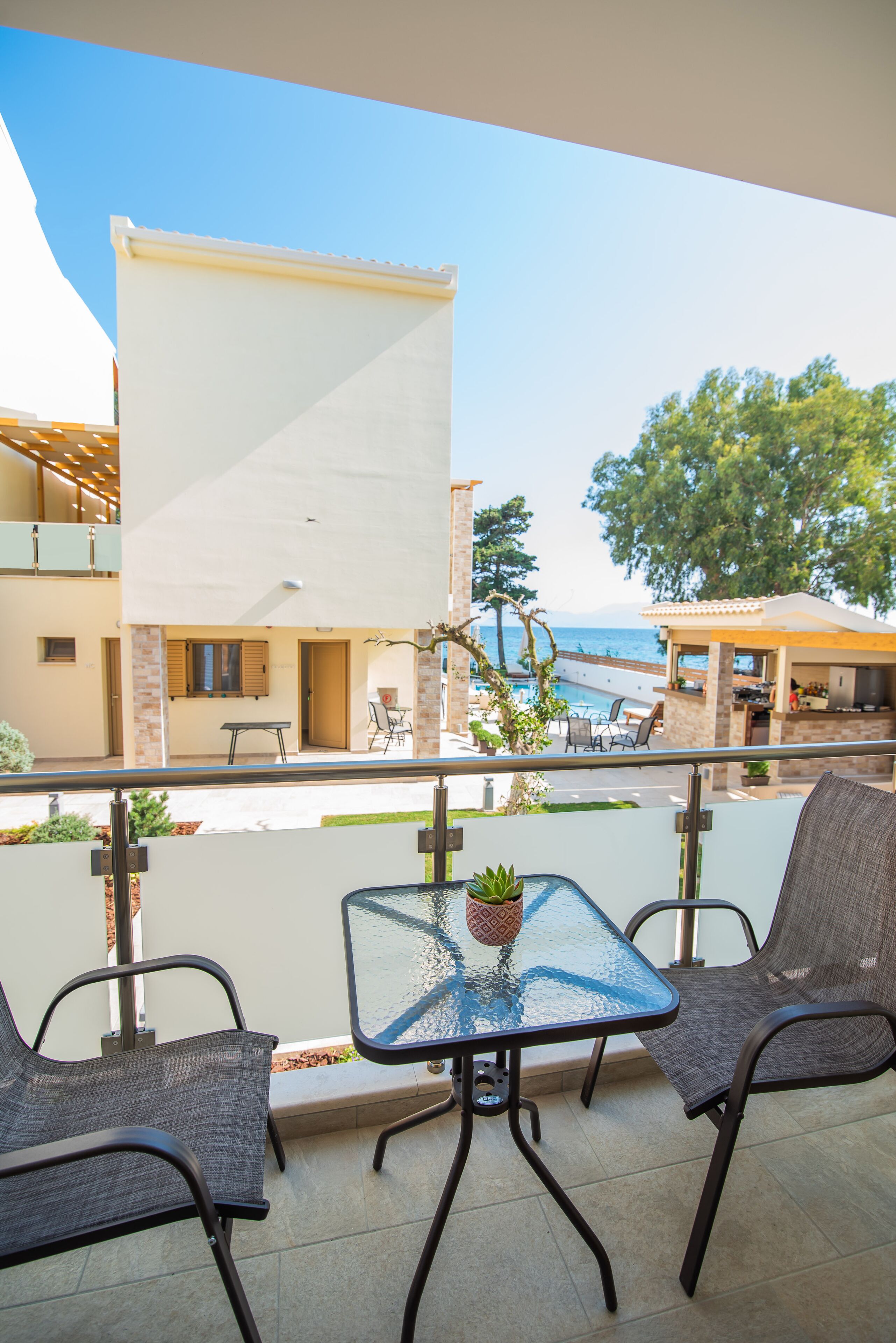 A balcony with a glass table and two chairs, overlooking a pool and a building with a beach view.