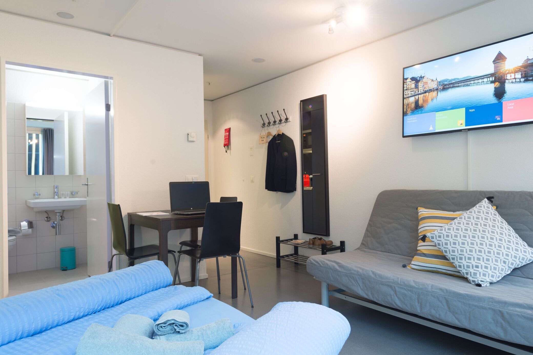 A hotel room with a bed, a desk with a computer, a mirror, a TV, and a window with a cityscape view.