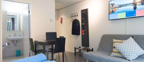 A hotel room with a bed, a desk with a computer, a mirror, a TV, and a window with a cityscape view.