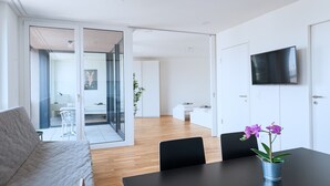A modern, well-lit room with a sofa, a dining table with chairs, a TV mounted on the wall, and a sliding glass door leading to another room. - Hitrental Basel Apartments (Basel)