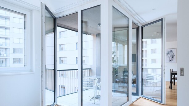 A modern apartment with large sliding glass doors leading to a balcony, a wooden floor, and a view of a building across the street. - Hitrental Basel Apartments (Basel)