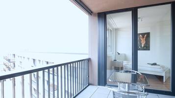 A balcony with a metal railing, a glass door leading to a room with a bed, a small table, and a chair.
