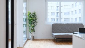 Ein modernes, minimalistisches Wohnzimmer mit großem Fenster, einem grauen Sofa, einem Topf mit Pflanze und einem spiegelnden Schwarzrahmen.
