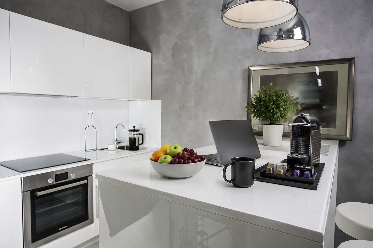 A modern kitchen with a built-in oven, a coffee maker, a laptop, and a bowl of fruit on the counter.