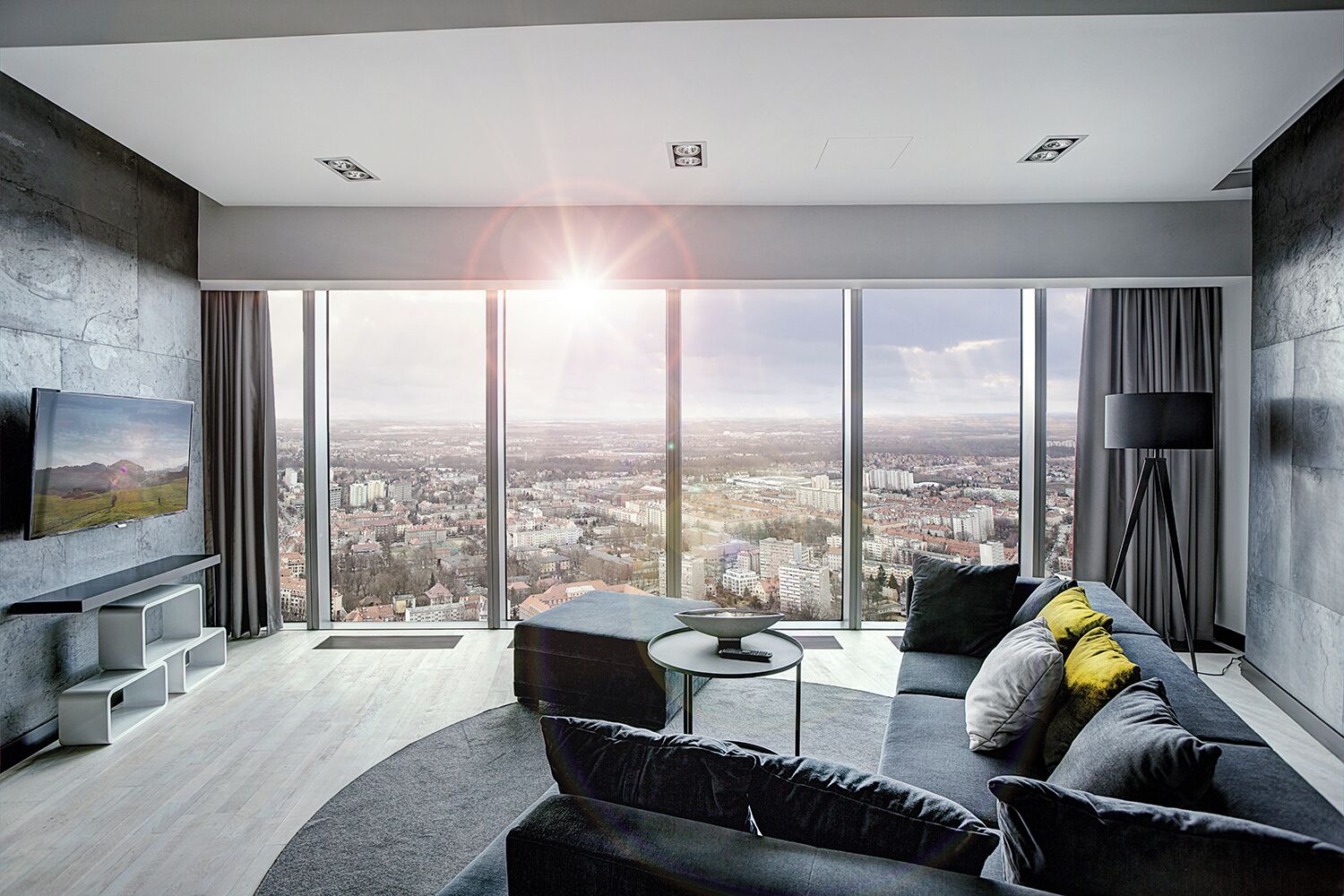 A modern living room with a large city view, a flat-screen TV mounted on the wall, a sofa with cushions, and a floor lamp.