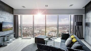 A modern living room with a large city view, a flat-screen TV mounted on the wall, a sofa with cushions, and a floor lamp.