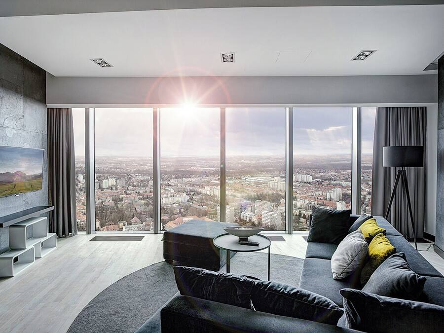 A modern living room with a large city view, a flat-screen TV mounted on the wall, a sofa with cushions, and a floor lamp.