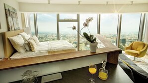 Een moderne slaapkamer met een groot bed, een nachtkastje met een plant, een gele fauteuil en uitzicht op de stad.