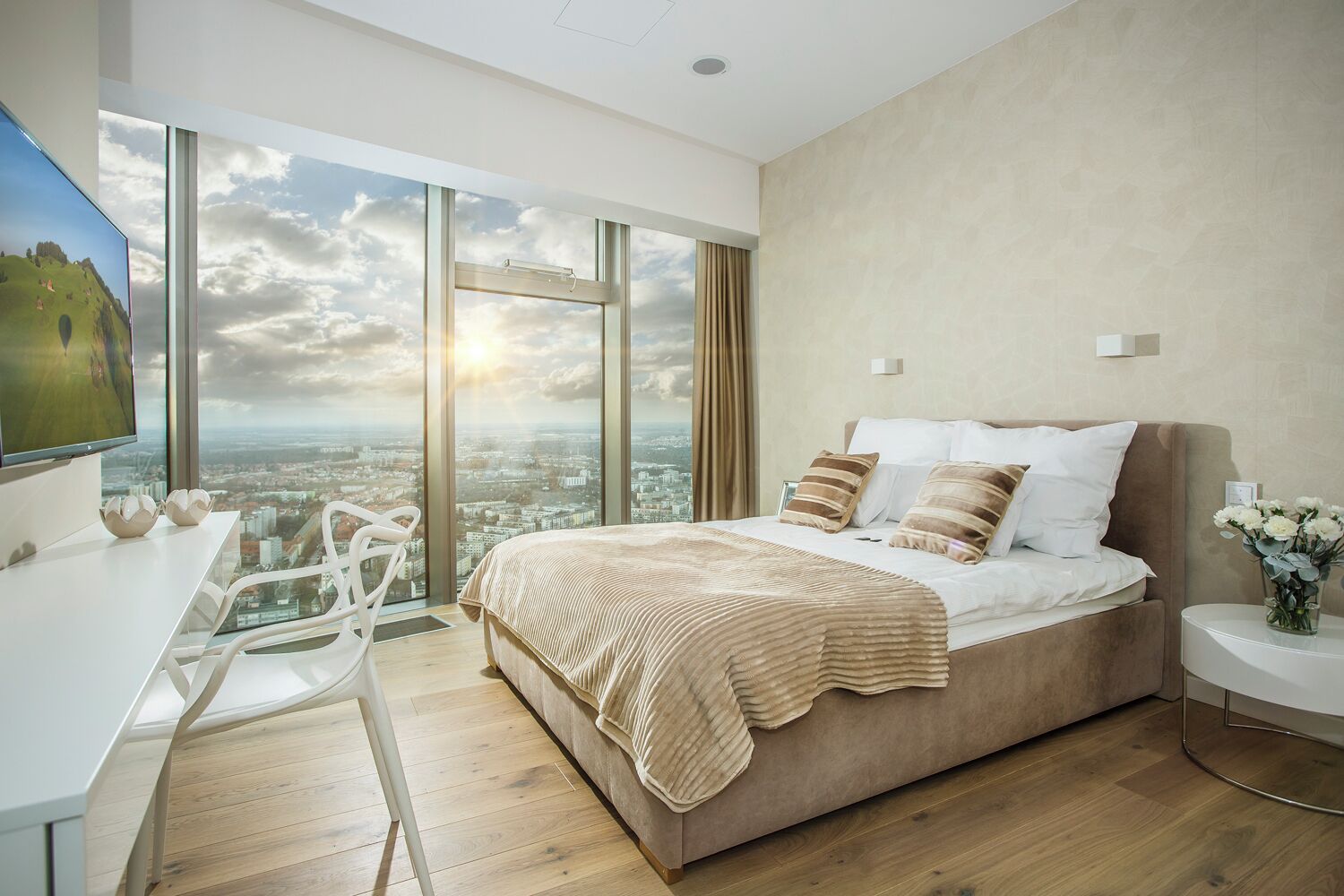 A modern bedroom with a large bed, a desk, and a view of a cityscape through large windows.