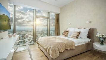 A modern bedroom with a large bed, a desk, and a view of a cityscape through large windows.