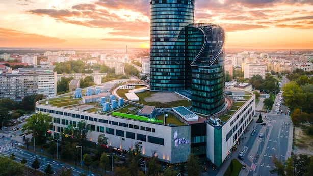 Een modern wolkenkrabber met een opvallend gebogen ontwerp, omgeven door een verzorgd stadsgebied met zichtbare wegen en andere gebouwen.