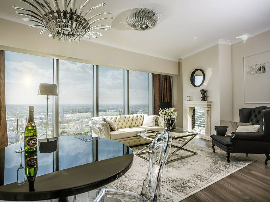 A spacious living room with a large window, a round glass table, a sofa, and a black leather armchair.
