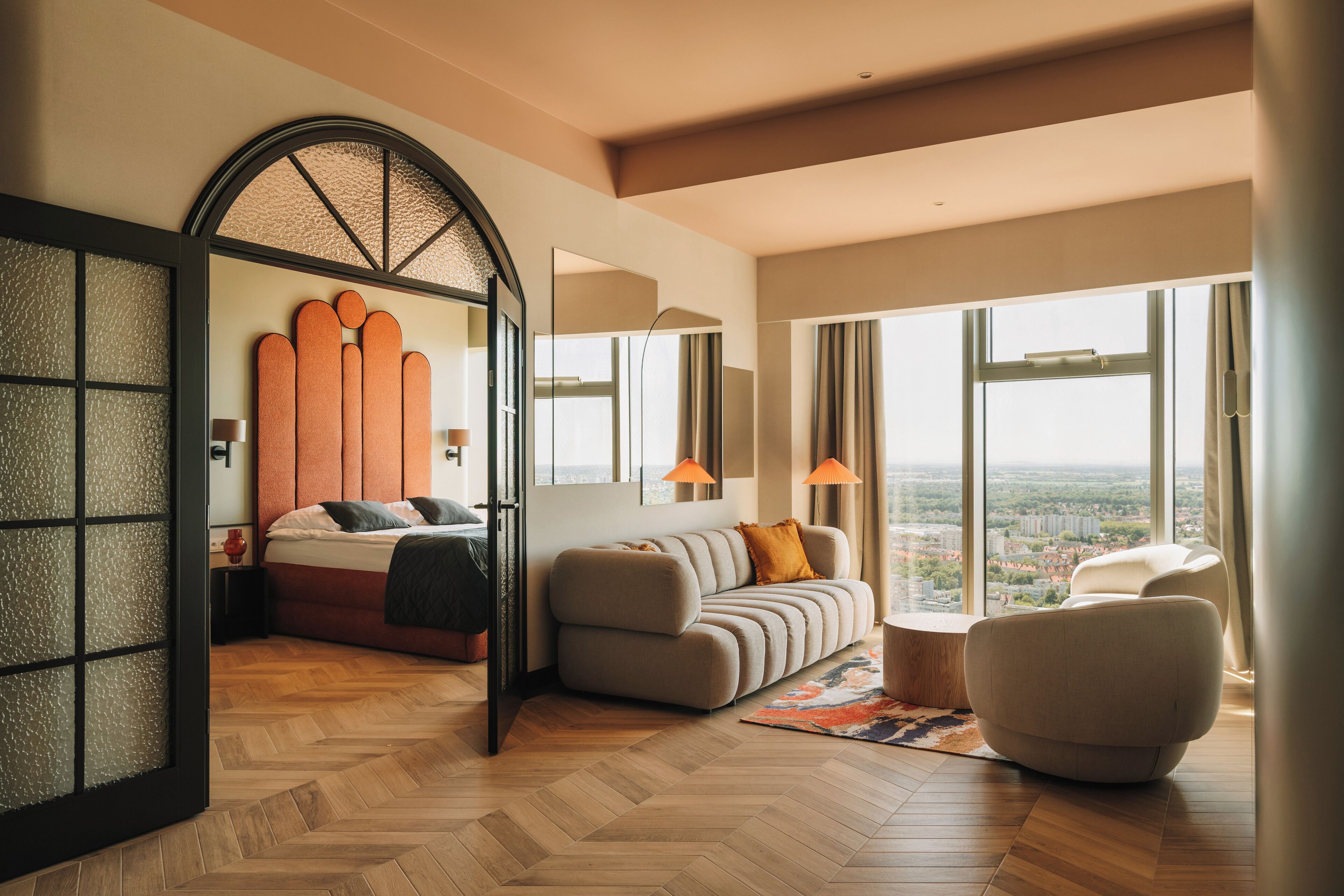 Una habitació d'hotel moderna amb un llit gran, un sofà, una cadira i vista a la ciutat a través de grans finestres.