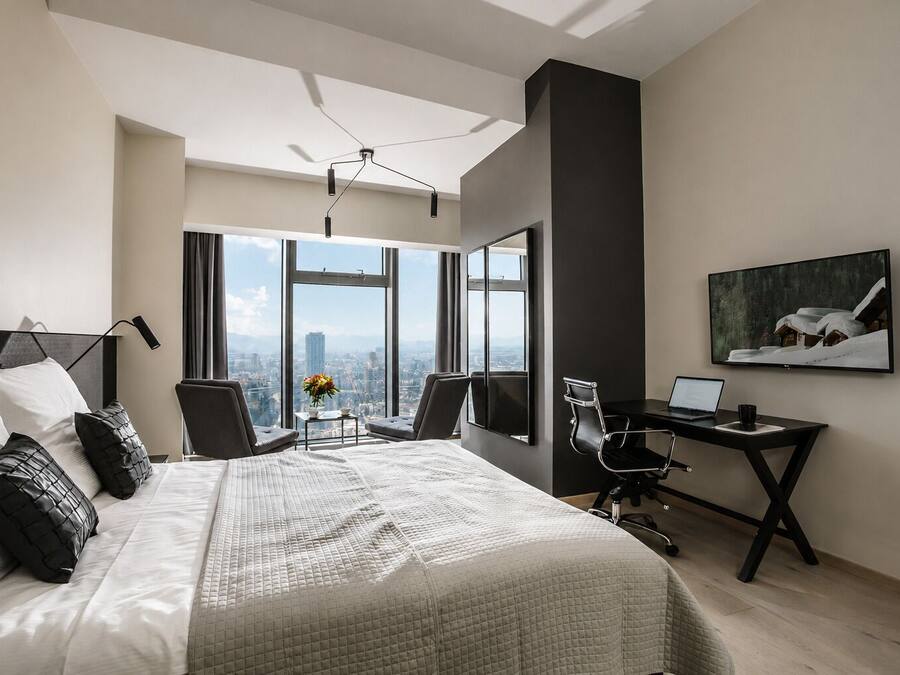 A modern hotel room with a large bed, a desk, a chair, a TV, and a city view.