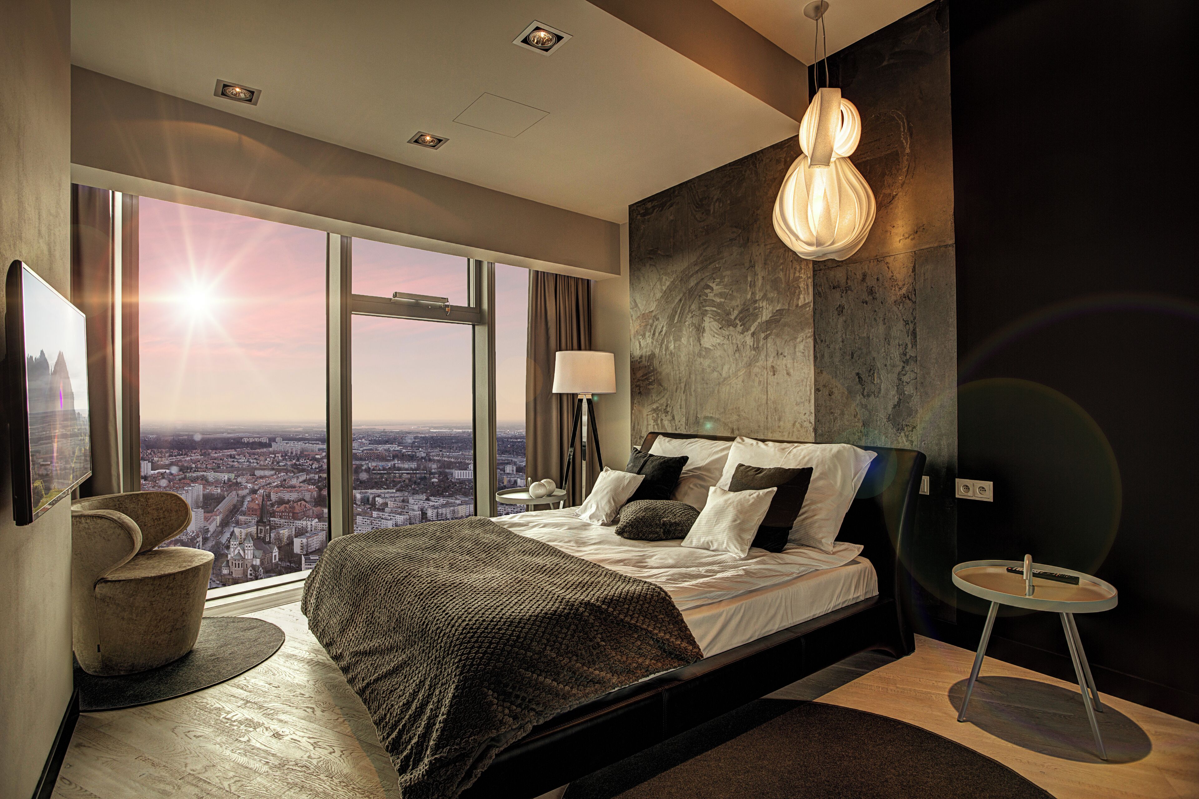 A modern bedroom with a large bed, a round side table, a floor lamp, and a view of the city through large windows.