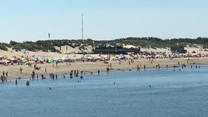 Una playa con gente nadando y tomando el sol. Hay tiendas de campaña y un escenario montado sobre la arena.